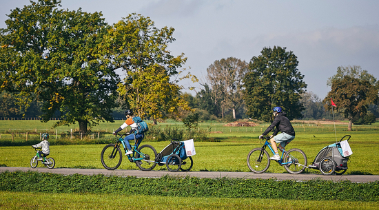 Das Bild zeigt eine Familie beim Fahrradausflug. Die Eltern fahren Fahrräder mit angehängtem Kiki, davor fährt ein kleines Kind. Im Hintergrund sind grüne Wiesen und Bäume zu sehen. 