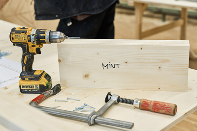 Auf dem Bild sieht man ein Stück Holz mit der Beschriftung "MINT" und ein Akkuschrauber und eine Holzbearbeitungsklemme mit Nägeln. 