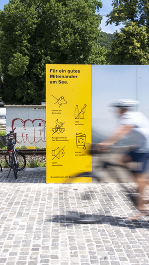 Auf dem Bild ist ein Fahrradfahrer vor einem gelben Hinweisplakat zu sehen mit den Regeln zum Verhalten am Seeufer.