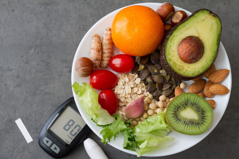 Das Bild zeigt einen Teller mit gesundem Essen, darunter eine Avocado, eine Kiwi, Erdbeeren, Nüsse und eine Orange. Daneben liegt ein Blutzuckermessgerät.