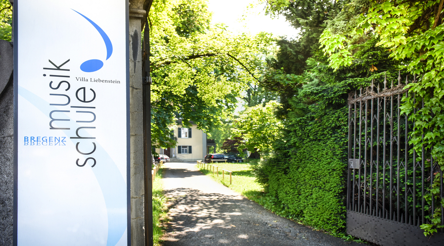 Das Bild zeigt das geöffnete gusseiserne Eingangstor zur Musikschule Bregenz. Dahinter ist das Gebäude der Villa Liebenstein, in dem die Musikschule untergebracht ist, zu sehen. Links im Bild ist ein Schild mit der Aufschrift "Villa Liebenstein" und dem Logo der Musikschule an einer Mauer angebracht. 