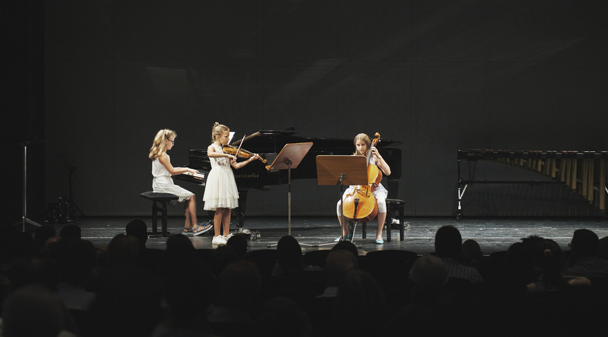 Auf dem Bild sind drei Schülerinnen der Musikschule Bregenz auf der Bühne zu sehen. Sie spielen Klavier, Geige und Cello.