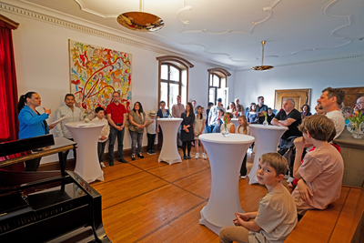 Das Bild zeigt den Empfang für die Gewinner:innen des "prima la musica"-Landeswettbewerbes, die im Salon der Musikschule Bregenz um Stehtisch stehen, während sie links im Bild Musikschuldirektorin Bettina Wechselberger begrüßt.