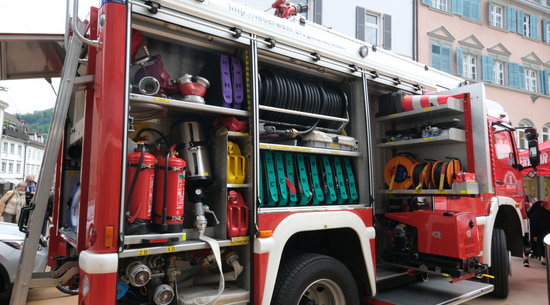 Das Bild zeigt ein Feuerwehrauto von der Seite, bei dem der Verstauraum geöffnet wurde und somit die geladenen Gerätschaften wie Schlauch, Sauerstoffflaschen und Ähnliches sichtbar sind. 