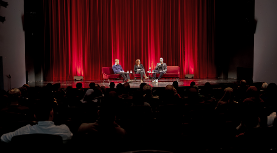 Auf rot-schwarzem Hintergrund sind drei Personen auf einer Bühne zu sehen
