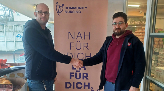 Das Bild zeigt Community Nurse Peter Köb und Mehmet Tag aus der Achsiedlung. Beide schütteln sich die Hände vor einem rosafarbenen Roll-Up mit der Aufschrift "Nah für Dich. Da für Dich". Oben ist das Logo von Community Nursing zu sehen. 