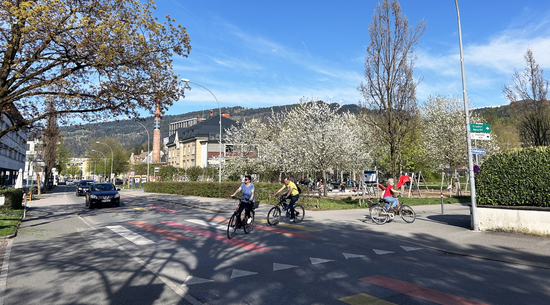 Auf dem Bild ist die Mariahilfstraße zu sehen. Auf dem Zebrastreifen sind drei Fahrradfahrende zu sehen und ein Auto. 