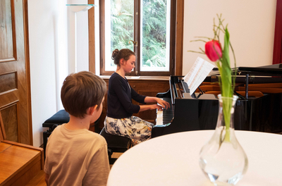 Das Bild zeigt eine Gewinnerin des "prima la musica"-Landeswettbewerbes, die ein Musikstück auf dem Klavier zum Besten gibt. Im Bildvordergrund sind ein Stehtisch mit einer Blume sowie ein kleiner Bub zu sehen, der ihr zuhört. Sie sitzt in einem Raum an einem Klavier vor einem Fenster.