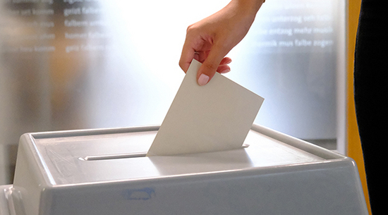 Auf dem Foto ist eine Hand mit einem Kuvert und einer Wahlurne zu sehen.