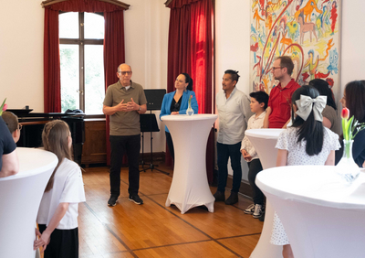 Das Bild zeigt den Empfang für die Gewinner:innen des "prima la musica"-Landeswettbewerbes, die im Salon der Musikschule Bregenz um Stehtisch stehen, während sie Kulturstadtrat Michael Rauth begrüßt.