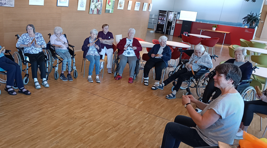 Auf dem Bild sind mehrere Seniorinnen zu sehen, die im Kreis auf Stühlen sitzen. Rechts vorne im Bild sind jüngere Personen, eine davon mit Klangstäben.
