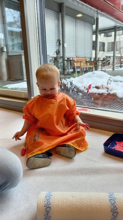 Auf dem Bild ist ein lachendes Kleinkind auf dem Boden sitzen und im Malkittel gekleidet zu sehen. 