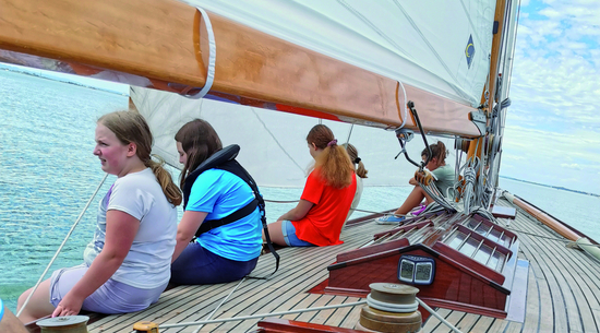 Das Bild zeigt mehrere Jugendliche, die beim Segeln auf dem Deck des Segelbootes sitzen und ins Wasser blicken. 