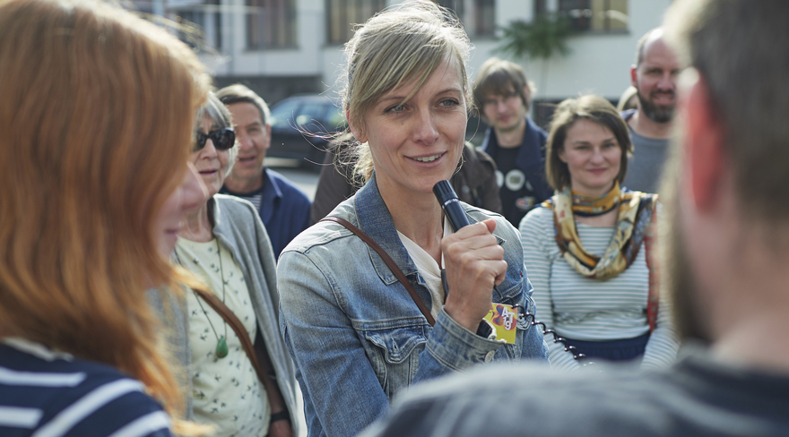 Diese Abbildung zeigt eine Frau, welche vor anderen Teilnehmer:innen des Stadtspazierganges eine Ansprache hält.