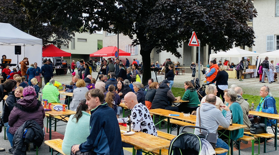 Das Bild zeigt die Mariahilfstraße sowie den Kirchenvorplatz, auf denen viele Menschen fürs Vorklöschtner Fescht versammelt sind. Sie sitzen an Bierbänken auf der Straße oder stehen auf dem Kirchenvorplatz an verschiedenen Ständen.