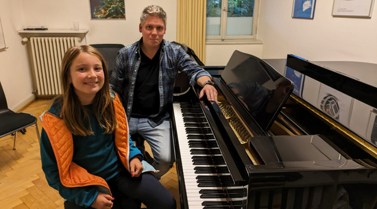 Auf dem Bild ist Kinderreporterin Larissa (vorne) mit Klavierlehrer Fabian Utz zu sehen, die auf einer Klavierbank vor einem schwarzen Klavier sitzen. 
