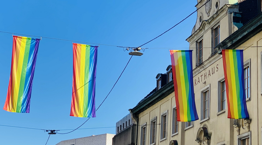 In dieser Abbildung sind bunte Pride-Flaggen zu sehen, welche über der Straße vor dem Rathaus hängen.