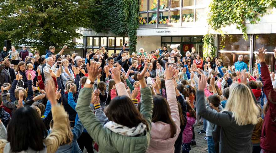Diese Abbildung zeigt einige Bürger:innen, welche sich vor der Schule Stadt versammelt haben und ihre Hände in die Höhe halten.