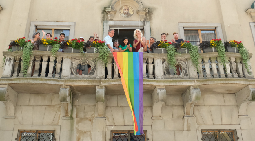 Diese Abbildung zeigt Markus Linhart mit anderen Personen auf dem Balkon des Rathauses. Gemeinsam bringen sie die Regenbogenfahne an. 