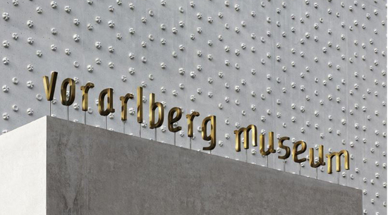 Das Bild zeigt die Buchstabeninstallation über dem Eingang der vorarlberg museum, bei der goldene Buchstaben den Schriftzug "vorarlberg museum" bilden. Dahinter die weiße Fassade des Museums. 