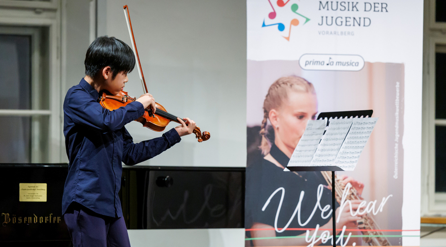 Das Bild zeigt einen Jungen in blauem Anzug beim Violine spielen, im Hintergrund stehen ein Flügel und ein Roll-Up zu "prima la musica".