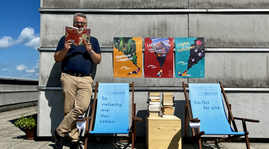 Diese Abbildung zeigt einen Mann, welcher ein Buch liest. Neben ihm sind zwei Stühle, Bücher und Plakate zu sehen.