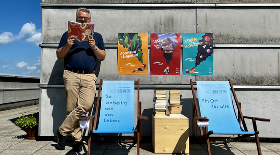 Diese Abbildung zeigt einen Mann, welcher ein Buch liest. Neben ihm sind zwei Stühle, Bücher und Plakate zu sehen.