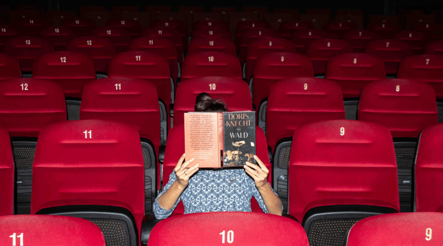 Das Bild zeigt eine Person, die auf einem Kinosessel in einem leeren Kinosaal sitzt und ein Buch vor ihr Gesicht hält und liest.