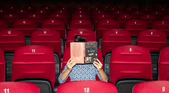 Das Bild zeigt eine Person, die auf einem Kinosessel in einem leeren Kinosaal sitzt und ein Buch vor ihr Gesicht hält und liest.