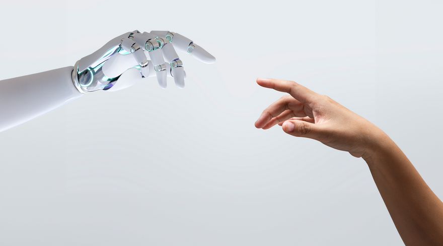 Das Bild zeigt eine künstliche Roboterhand auf der linken Seite und eine menschliche Hand auf der rechten Seite, die beide den Zeigefinger ausstrecken und sich beinahe berühren. Der Hintergrund ist einfärbig grau.