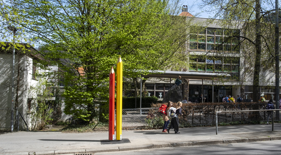 Das Bild zeigt zwei überdimensionale Buntstifte, die auf dem Gehsteig vor einer Schule platziert sind. In Hintergrund sind die Schule und Bäume zu sehen, rechts im Bild laufen zwei Schülerinnen Richtung Schule. 