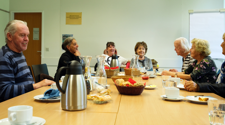 Mehrere Senior:innen sitzen beim Dienstagsfrühstück an einem Tisch und frühstücken zusammen, während sie Gespräche führen. 