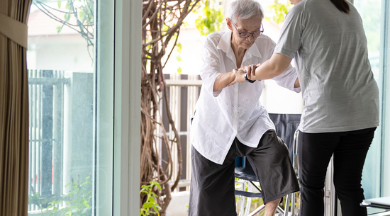 Das Bild zeigt eine alte Dame, der beim Betreten einer Wohnung über eine Stufe von der Terrasse aus von einer jungen Pflegerin geholfen wird. 