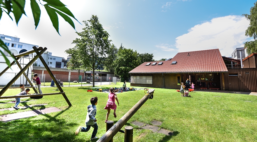 Das Bild zeigt im Hintergrund einen Kindergarten, davor eine große grüne Wiese mit einem kleinen Spielplatz, auf dem Schaukeln und Spielgerät zu sehen sind. Im Bild rennen ein paar Kinder auf der Wiese in Richtung Kindergarten.